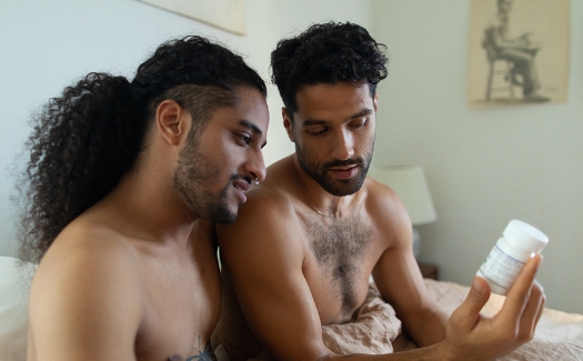 Two men in bed together looking at a medication bottle